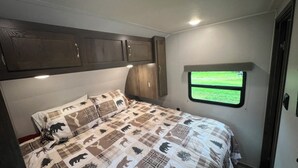 A bedroom with a bed, a window, and wooden cabinets. - EastToWest Glamping camper - Site #15 (Klamath)