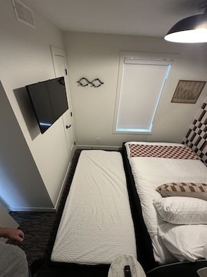 A small hotel room with two beds, a TV, a window with blinds, and a ceiling vent. - The Station 810 | Downtown | Rooftop Hot Tub (Canmore)