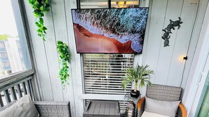 A balcony with a sofa, a small table, a potted plant, a TV mounted above the window, and a patterned rug. -  Scenic Golf Course View Retreat - UPDATED 2BR Condo-Close to Beach (North Myrtle Beach)