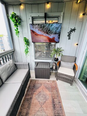 Ein Balkon mit einer Couch, einem kleinen Tisch, einem Blumentopf, einem über dem Fenster montierten Fernseher und einem gemusterten Teppich.