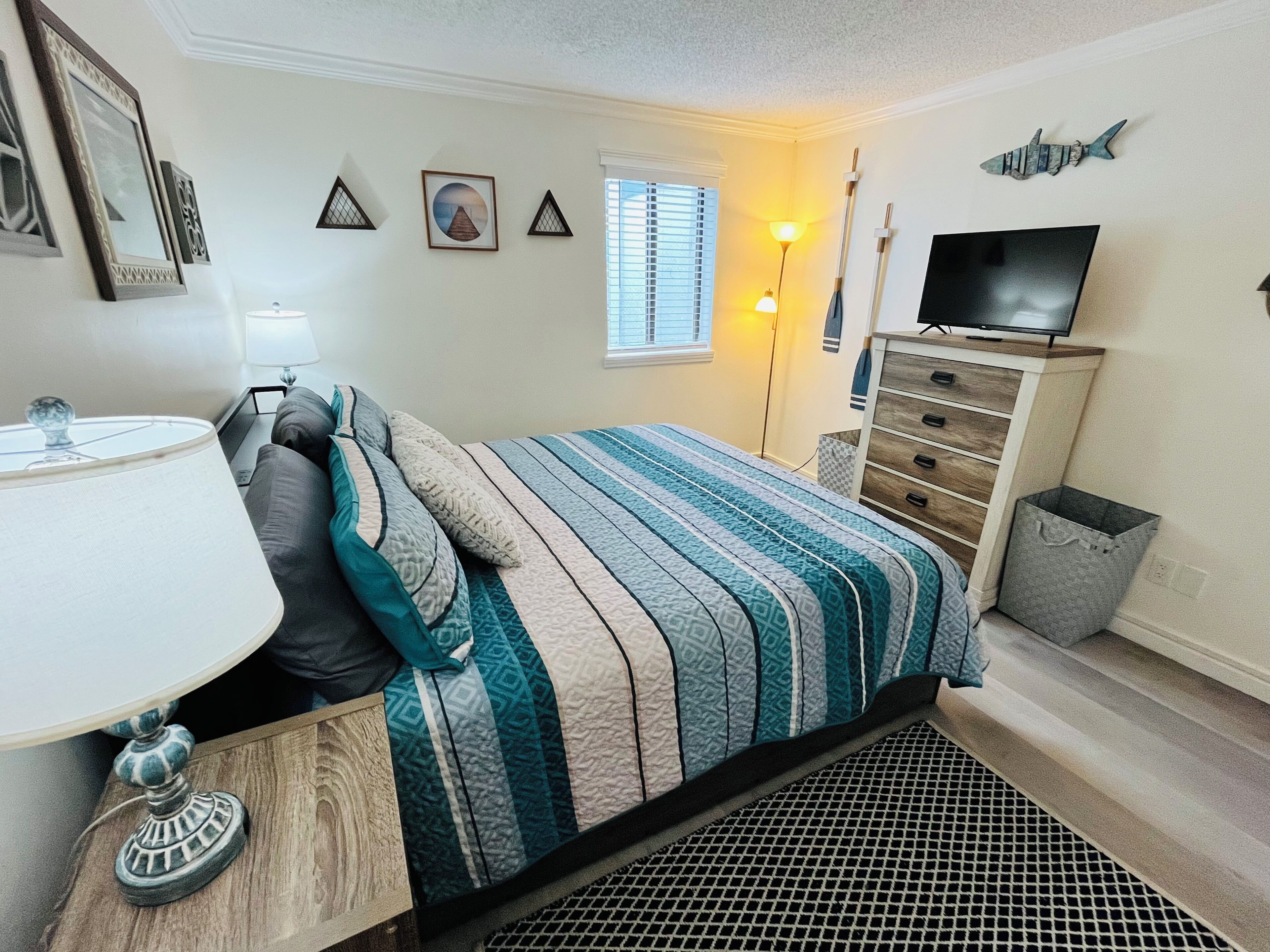 Une chambre à coucher comprenant un lit, une commode, un téléviseur, une lampe et une fenêtre avec des stores.