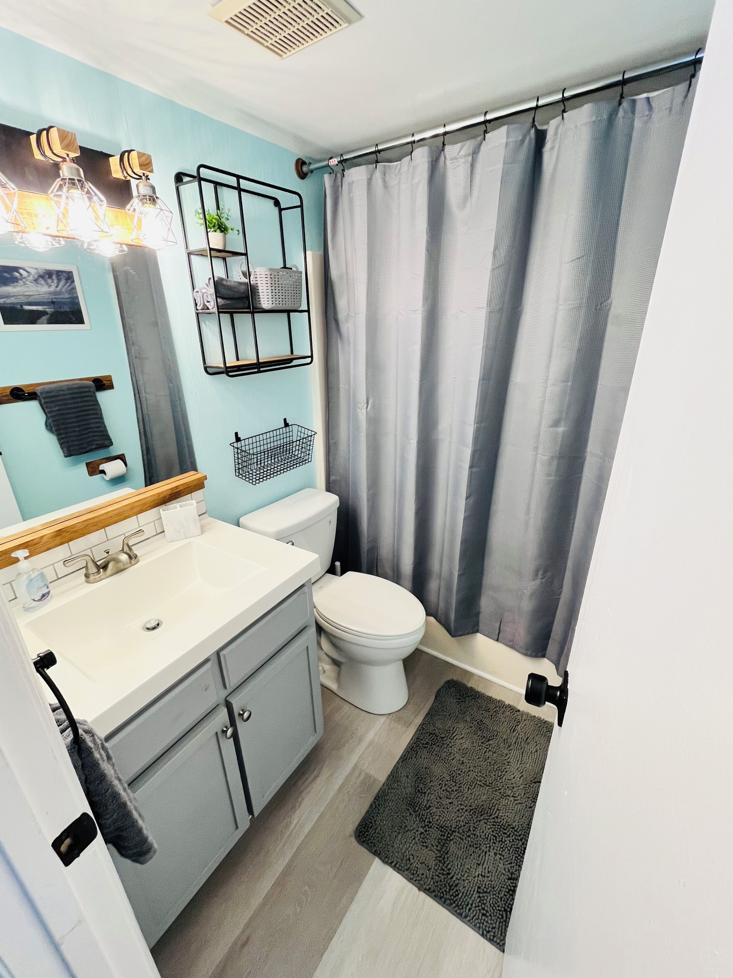 A bathroom with a white sink, toilet, and shower curtain. There is a towel rack, a small shelf with baskets, and a picture on the wall.