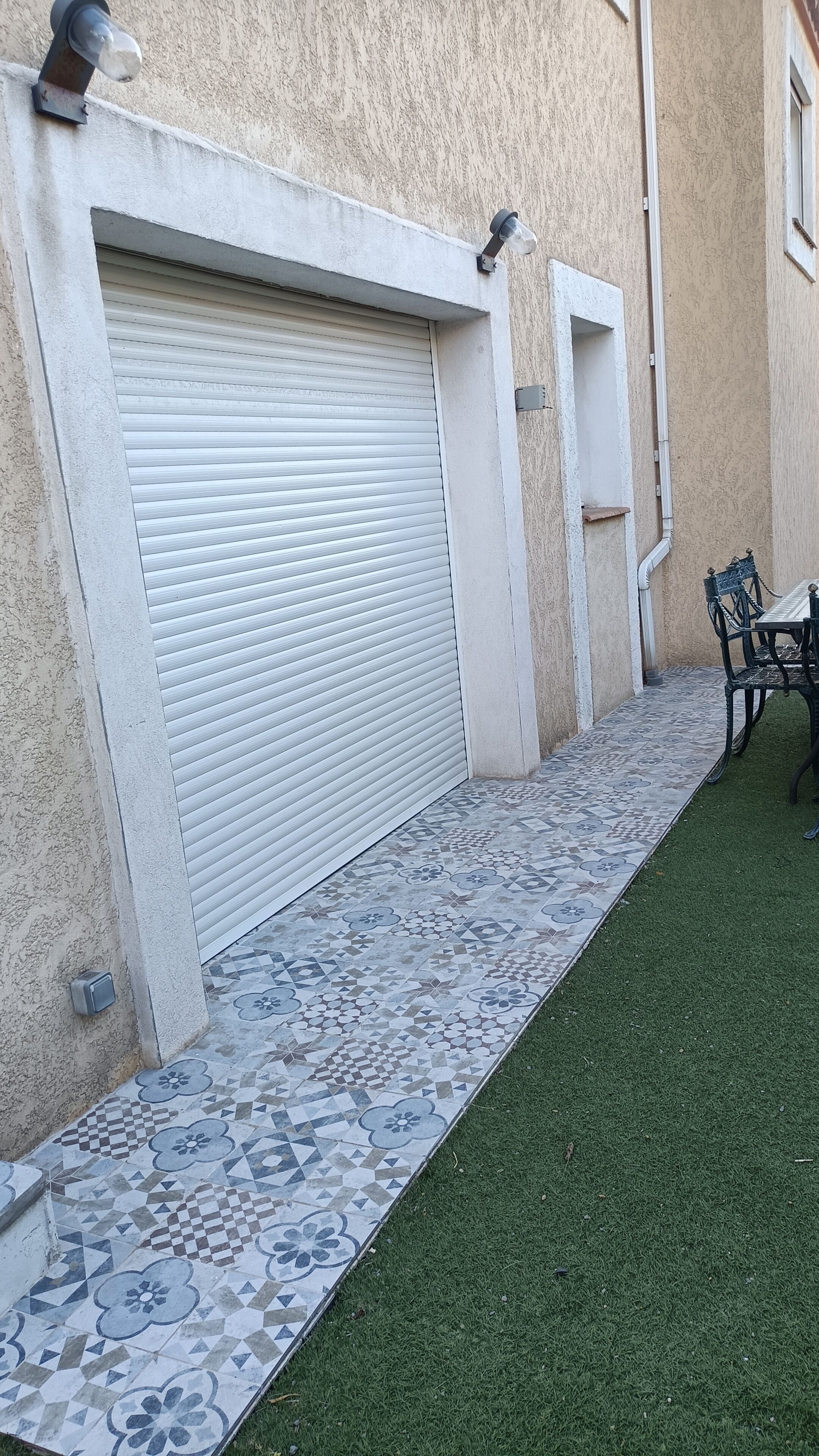 A building with a closed garage door, a tiled pathway, and a small outdoor seating area with a table and chairs.