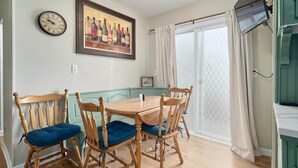 A dining area with a wooden table and chairs, a clock, a framed wine bottle artwork, and a sliding glass door.