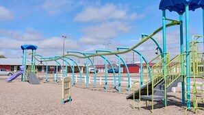 A playground with slides and climbing structures, surrounded by a fence and buildings in the background.