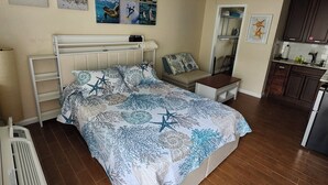 A bedroom with a bed, a ceiling fan, a nightstand, a wall-mounted heater, and a kitchen area with cabinets and a refrigerator.