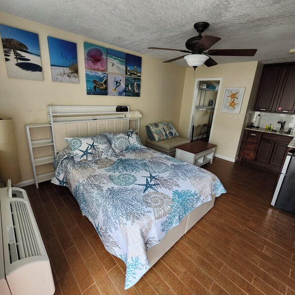 A bedroom with a bed, a ceiling fan, a nightstand, a wall-mounted heater, and a kitchen area with cabinets and a refrigerator.