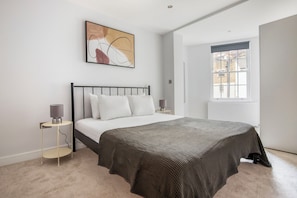 Ein Schlafzimmer mit Bett, Nachttisch, Lampe, Fenster und einem Bild an der Wand.