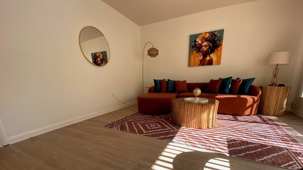 A living room with a wooden floor, a round coffee table, a sofa with cushions, a painting on the wall, a floor lamp, and a mirror.