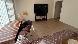 A modern living room with a wooden floor, a flat-screen TV on a stand, a dining table with chairs, and a decorative vase with pampas grass.
