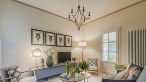 A living room with a sofa, armchair, coffee table, TV, framed artwork, chandelier, and a window with blinds. - Charming Lucca Villa With Garden (Lucca)