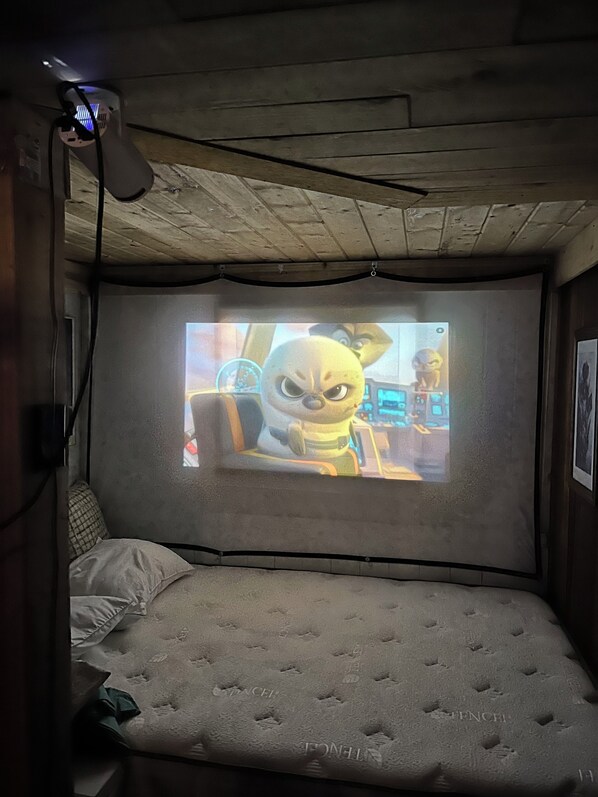 A room with a bed, a projector screen displaying an animated character, and a ceiling with visible beams.