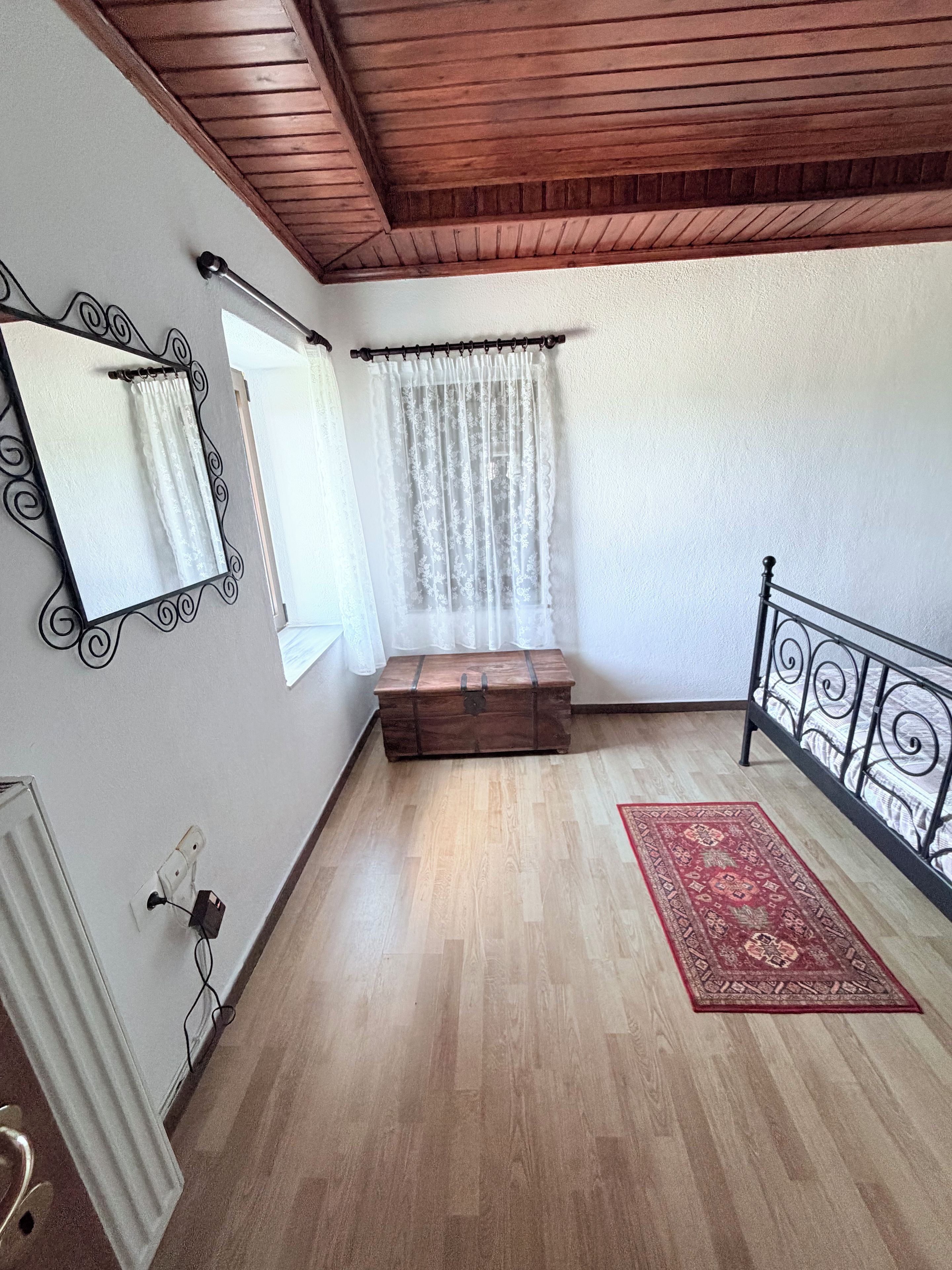 A room with a wooden ceiling, a bed with metal headboard, a small wooden chest, a red patterned rug, and a window with sheer curtains.
