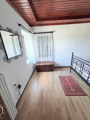 Una habitación con techo de madera, una cama con cabecera de metal, un pequeño cofre de madera, un tapete rojo con estampado y una ventana con cortinas traslúcidas.