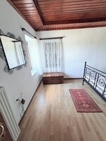 Una habitación con techo de madera, una cama con cabecera de metal, un pequeño cofre de madera, un tapete rojo con estampado y una ventana con cortinas traslúcidas.