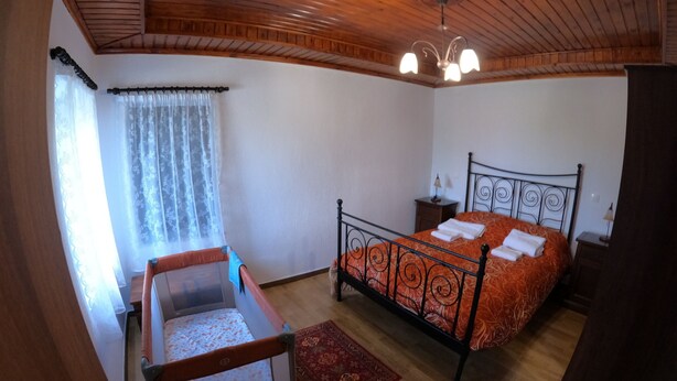 A bedroom with a wooden ceiling, a bed with an orange bedspread, a nightstand, a chair, and a window with sheer curtains.
