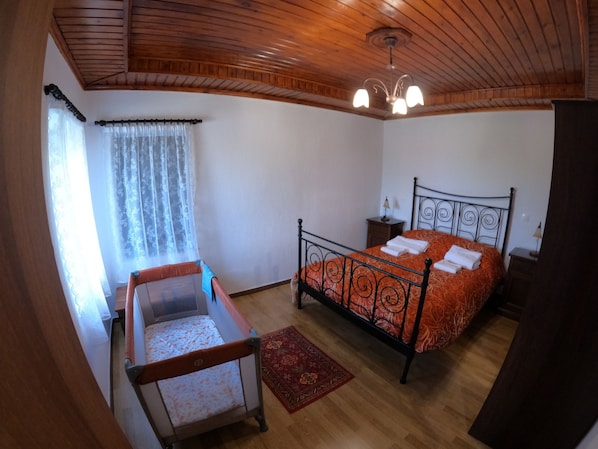 A bedroom with a wooden ceiling, a bed with an orange bedspread, a nightstand, a chair, and a window with sheer curtains. - PIALEIA  (Pyli)