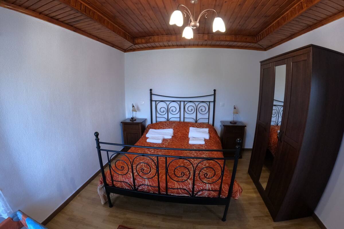 Une chambre avec un plafond en bois, un lit recouvert d’une couvre-lit à motifs, deux tables de chevet avec des lampes et une armoire.