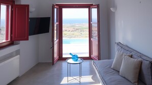 Et værelse med fjernsyn monteret på væggen, en sofa, et lille bord og havudsigt gennem åbne døre.