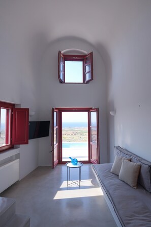A room with a TV mounted on the wall, a sofa, a small table, and a view of the sea through open doors. - Deluxe Villa with Plunge Pool in Oia (Oia)