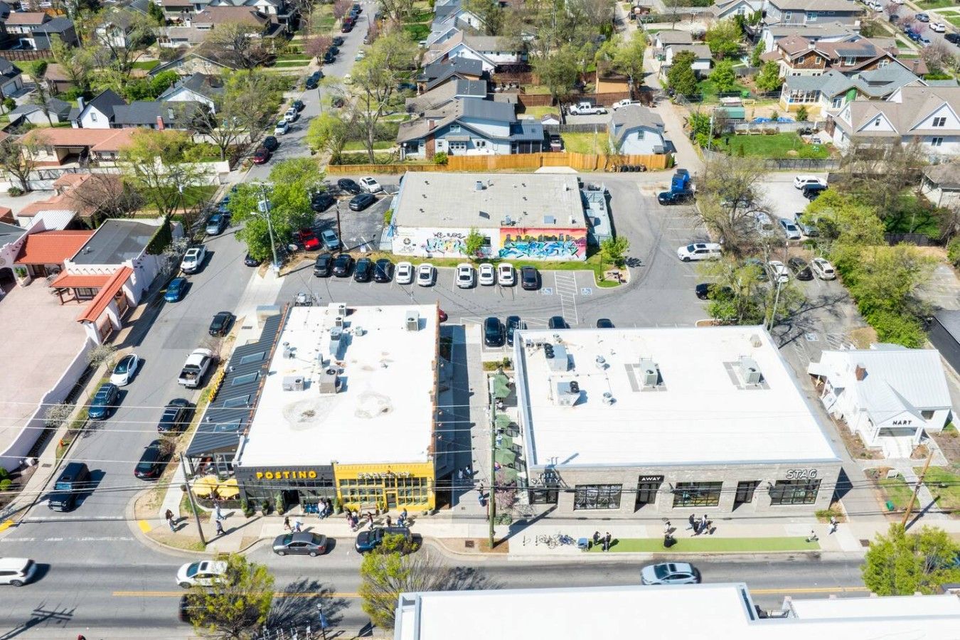 Vue aérienne d’une zone commerciale comprenant un parking, un bâtiment sur lequel on peut lire l’inscription « POSTINO », et une rue où circulent des voitures et des piétons.