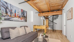 Uma sala de estar moderna com um sofá cinza, uma mesa de centro de madeira, um quadro e uma mesa de pebolim.