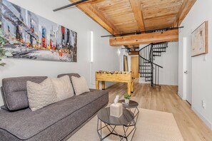 A modern living room with a gray sofa, a wooden coffee table, a painting, and a foosball table. - Downtown Two-Level Loft | Private Gym + Parking | Steps From Roger’s Place (Edmonton)
