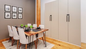 Uma área de refeições com mesa de madeira, cadeiras brancas, luminária pendente cinza e um guarda-roupa cinza com puxadores pretos.