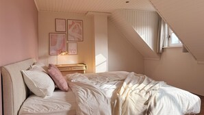 Een slaapkamer met een schuine hemel, een bed met witte beddengoed, een nachtkastje met een lamp en lijstjes met foto’s aan de muur.