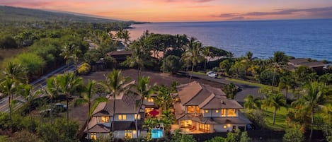 A large, multi-story house with a swimming pool, surrounded by palm trees and a view of the ocean.