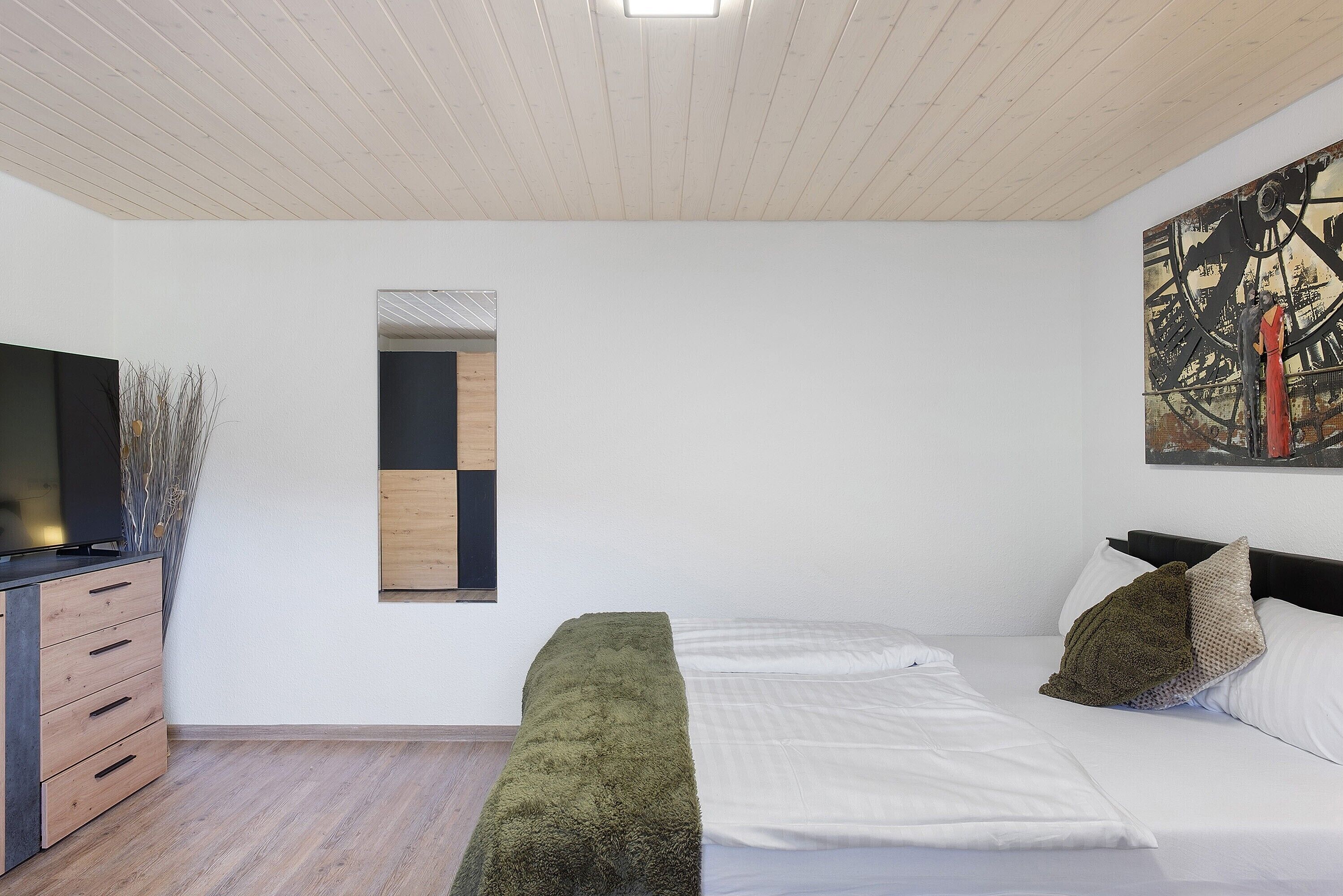 A modern bedroom with a bed, a dresser, a mirror, and a painting on the wall.