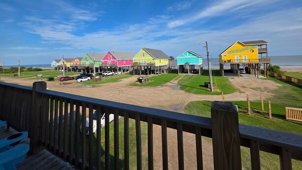 Una terraza de madera frente al mar, con edificios coloridos, una zona arenosa y vistas al océano.