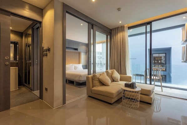 A modern hotel room with a sofa, a small round table, a bed, and a view of the city through large windows.