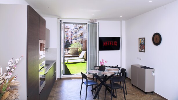 Een moderne keuken met een schuifdeur naar een balkon, een eethoek met een tafel en stoelen, en een aan de muur bevestigde televisie waarop 'NETFLIX' wordt weergegeven.
