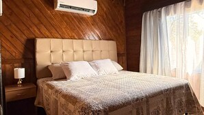 A bedroom with a wooden ceiling, a bed with a floral bedspread, a nightstand with a lamp, and a window with curtains. - Luxury cottage in front of the sea and lagoon of miami Tela (TELA)
