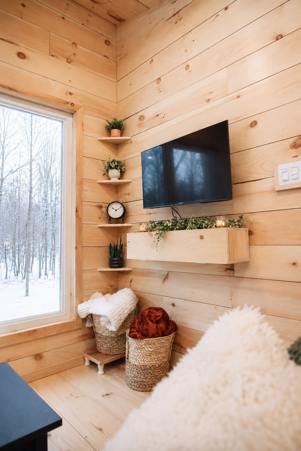 A cozy cabin interior with wooden walls, a mounted TV, shelves with plants, a clock, and a window with a snowy view.
