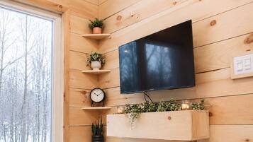 A cozy cabin interior with wooden walls, a mounted TV, shelves with plants, a clock, and a window with a snowy view.