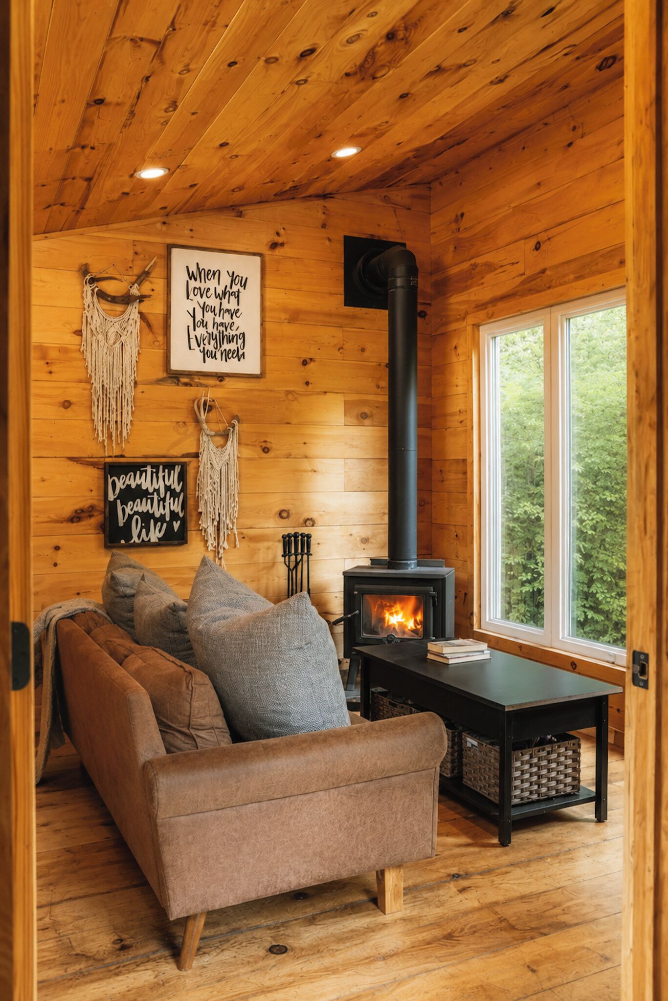 A cozy cabin interior with a wooden ceiling, a fireplace, a sofa, and a window overlooking greenery.