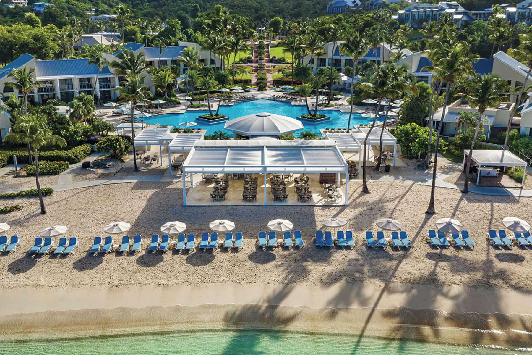Ein Luftbild eines Resorts mit einem zentralen Pool, mehreren Liegebereichen und einem Strand mit Sonnenliegen.