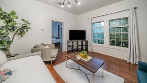 A modern living room with a flat-screen TV, a coffee table with decorative items, a grey sofa, and a large window with curtains. - Steps to Bishop Arts, Private Yard + Pet-Friendly (Dallas)