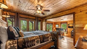 Interior de uma cabana de madeira com cama, escrivaninha e computador, com grandes janelas e um ventilador de teto.