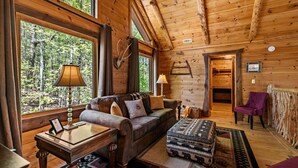 Interior de uma cabana de tronco com sofá, banquinho, luminárias e vista da floresta através de grandes janelas.