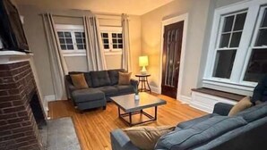 A living room with a brick fireplace, a grey sofa, a coffee table, and a lamp. - University Hill Entire 5 BR House -  Walk to SU campus, Upstate Hospital (Syracuse)