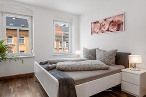 Ein Schlafzimmer mit einem Bett, Nachttischen, einer Lampe, einer Topfpflanze und einem Fenster.