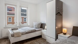 A bedroom with a bed, bedside table, lamp, wardrobe, and a window with a view of buildings. - [G2] Apartment for 6 People (75m²) in Gera (Gera)