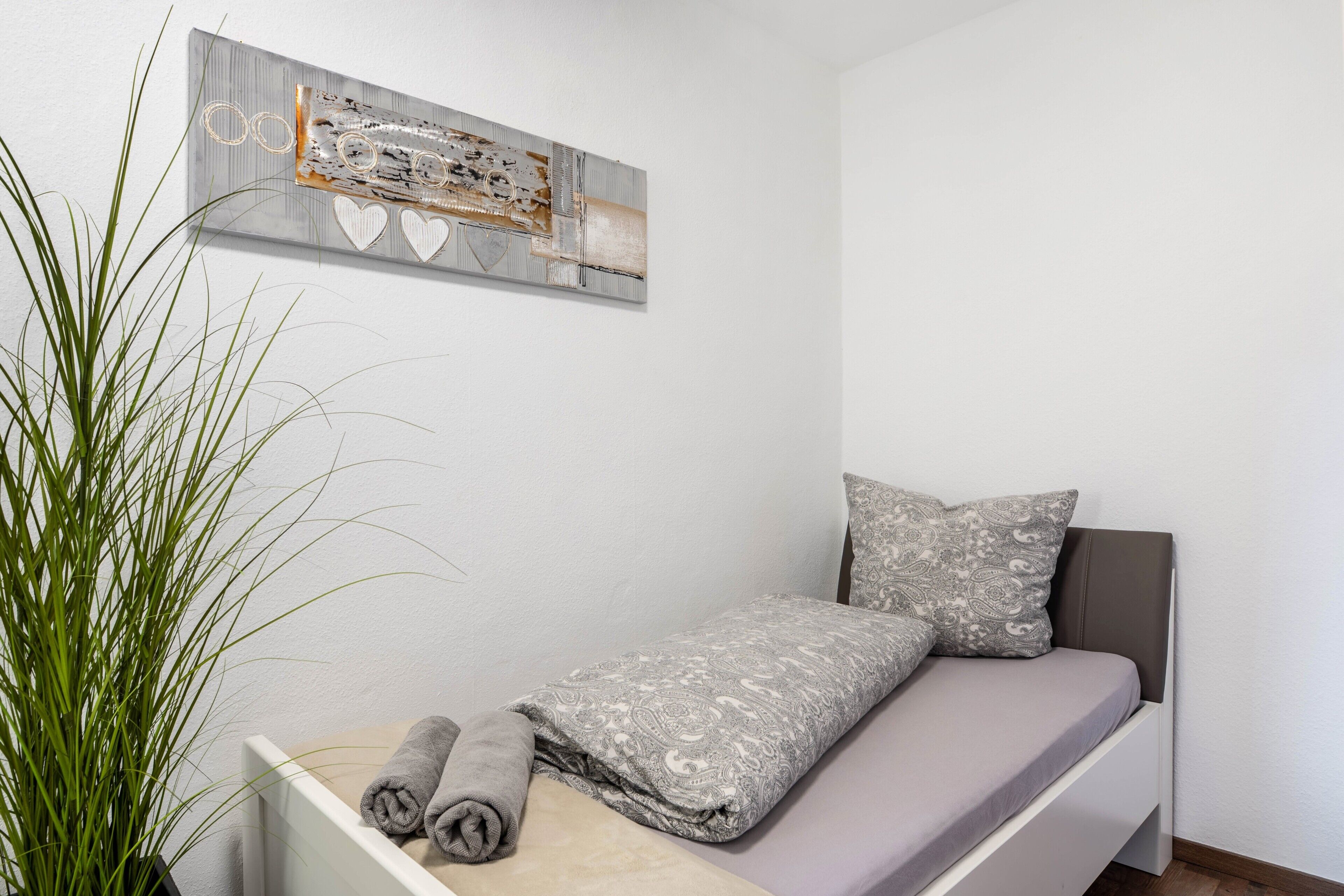 Ein schlicht eingerichtetes Schlafzimmer mit Bett, Kissen, einer Wanddekoration und einer Topfpflanze.
