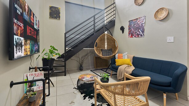 A living room with a blue sofa, wicker chairs, a staircase, and a wall-mounted TV displaying Netflix content. - Lofthaus Rosebank: walk to mall, safe & central. Family friendly 2BR with WIFI (Johannesburg)