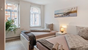 Une chambre avec deux lits, une table de chevet avec une lampe, une fenêtre avec des stores et un tableau au mur.