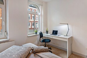 Ein Schlafzimmer mit Bett, Schreibtisch mit Laptop und Fenster mit Blick auf Gebäude.