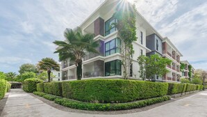 Een modern meerlaags gebouw met balkons, omgeven door verzorgde hagen en palmbomen.